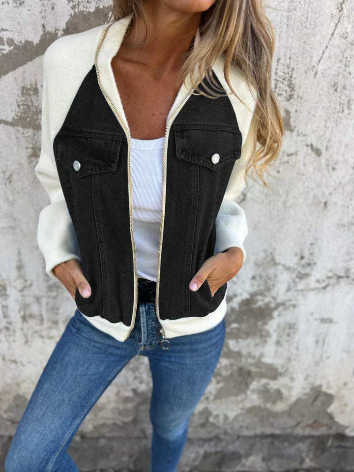 Frau in weißer Jacke mit schwarzen Taschen, Jeans, vor Betonwand.