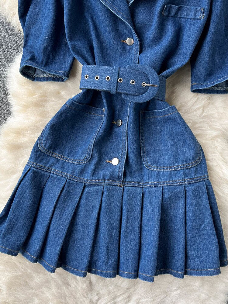 Blaues Jeanskleid mit Gürtel, Knöpfen und Faltenrock auf weißem Hintergrund.