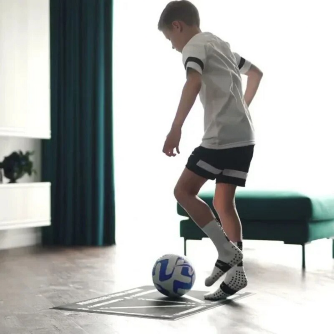 Junge spielt mit blau-weißem Fußball im Wohnzimmer, trägt Sportkleidung und Fußballschuhe.