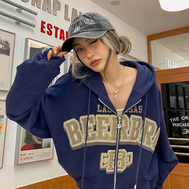 Frau in blauer Kapuzenjacke mit "BEERBR" Aufdruck und grauer Kappe, lässiger Streetwear-Look.