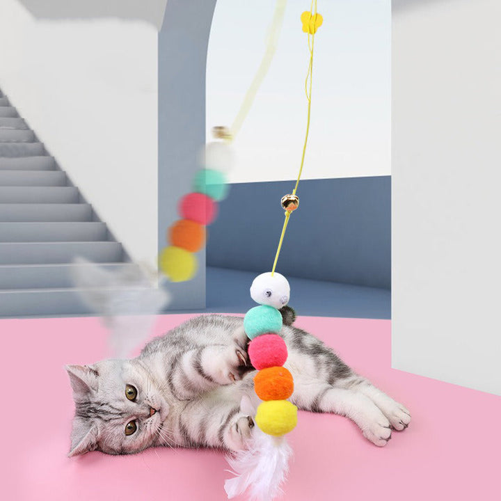 Graue Katze spielt mit buntem Katzenangelspielzeug, Pompons und Federn, auf rosa Boden.