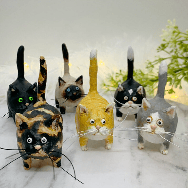 Sechs handgefertigte Katzenfiguren aus Ton, bunt bemalt, auf weißem Hintergrund mit Pflanzen.