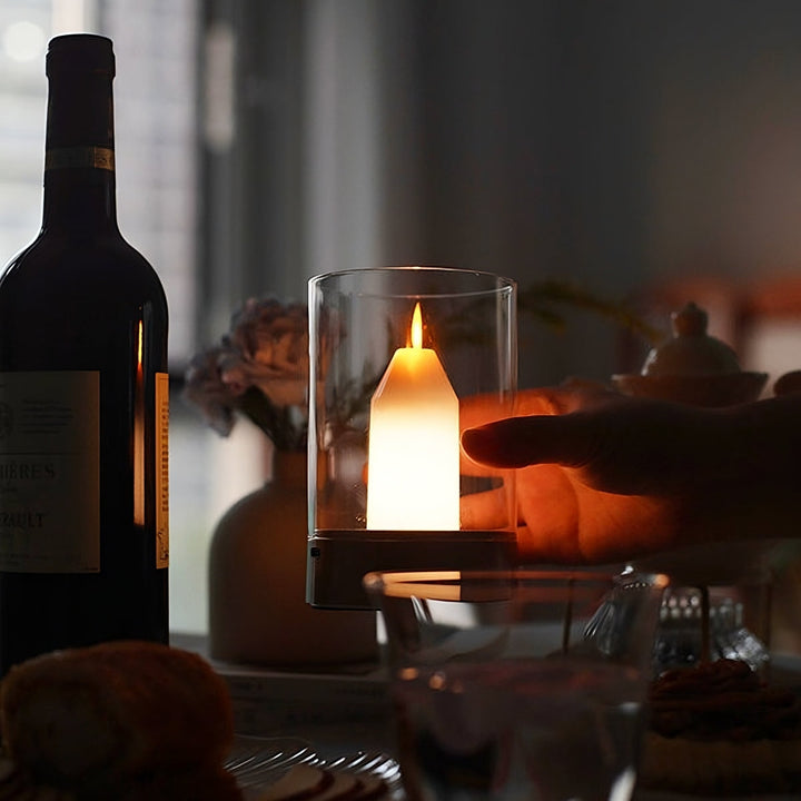Hand hält LED-Kerze in Glas, neben Weinflasche, auf gedecktem Tisch, stimmungsvolle Beleuchtung.