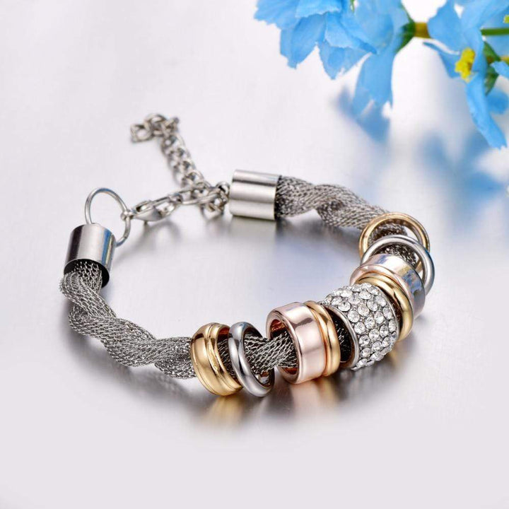 Verflochtenes Modearmband mit Metallringen und Strass, silberfarben, neben blauen Blumen.