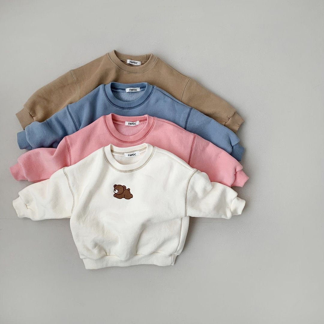 Vier Kinderpullover in Beige, Blau, Rosa und Weiß mit Bärenmotiv auf grauem Hintergrund.
