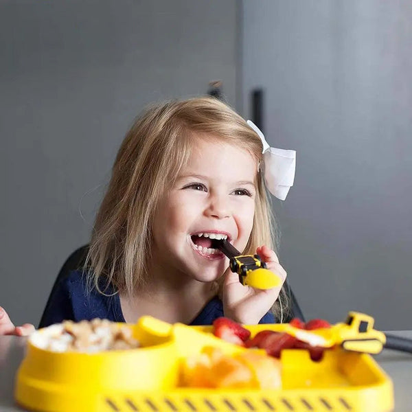Mädchen isst mit gelbem Bagger-Besteck, lächelnd, Kindergeschirr, spielerisches Essen.