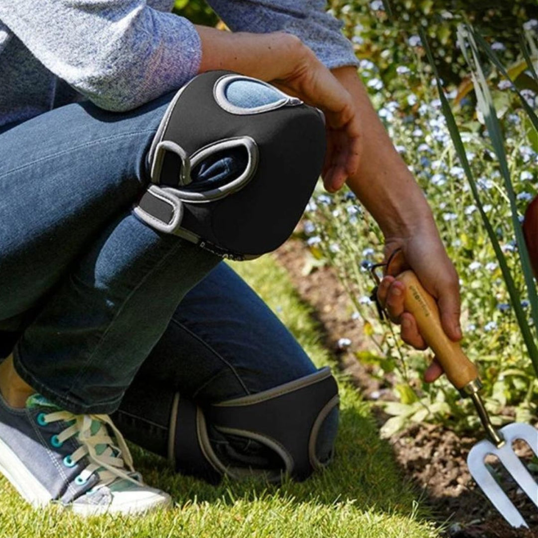 Person mit schwarzen Knieschonern beim Gärtnern, hält Gartenwerkzeug, Blumenbeet im Hintergrund.