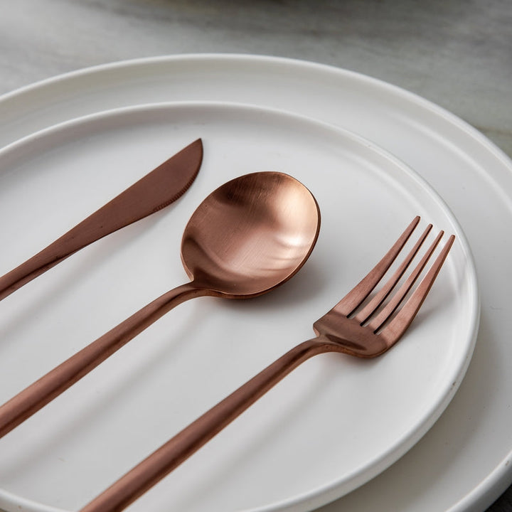 Kupferbesteck-Set auf weißem Teller, bestehend aus Messer, Gabel und Löffel, elegant und modern.