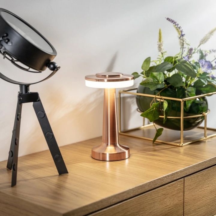 Kupferfarbene LED-Tischlampe auf Holzschrank neben schwarzem Scheinwerfer und Pflanze.