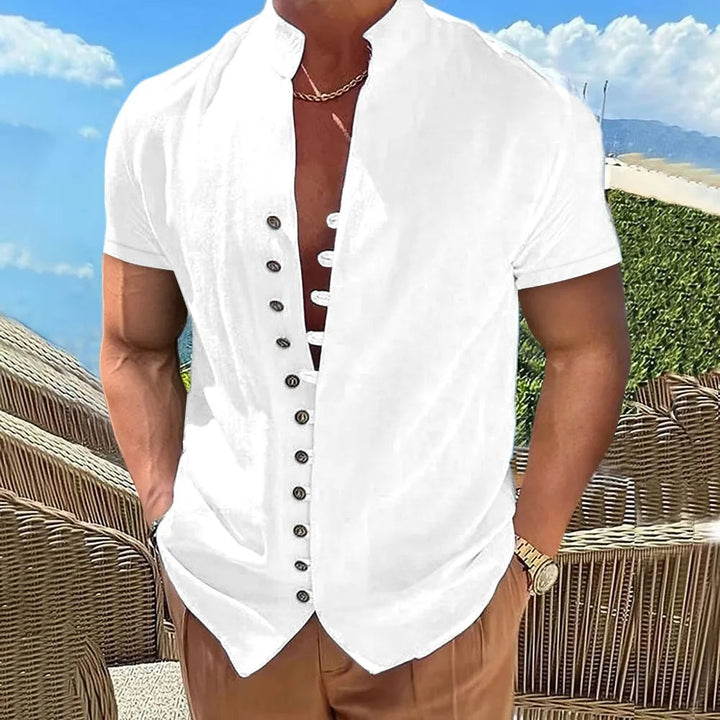 Mann in weißem Kurzarmhemd mit Knopfleiste, braune Hose, Strandhintergrund.