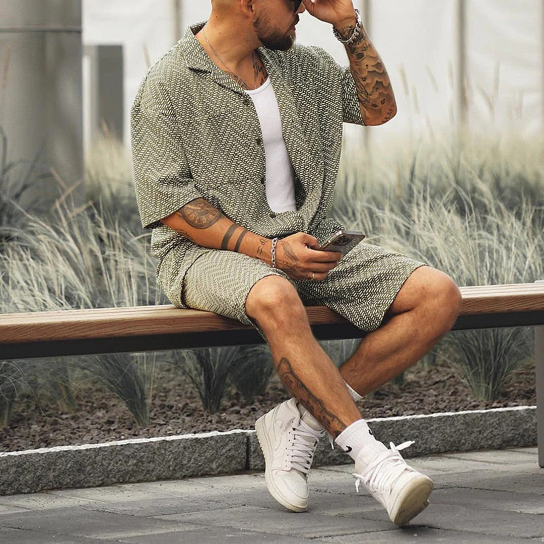 Mann in grünem Sommer-Set, sitzt auf Bank, trägt weiße Sneakers, hält Smartphone.