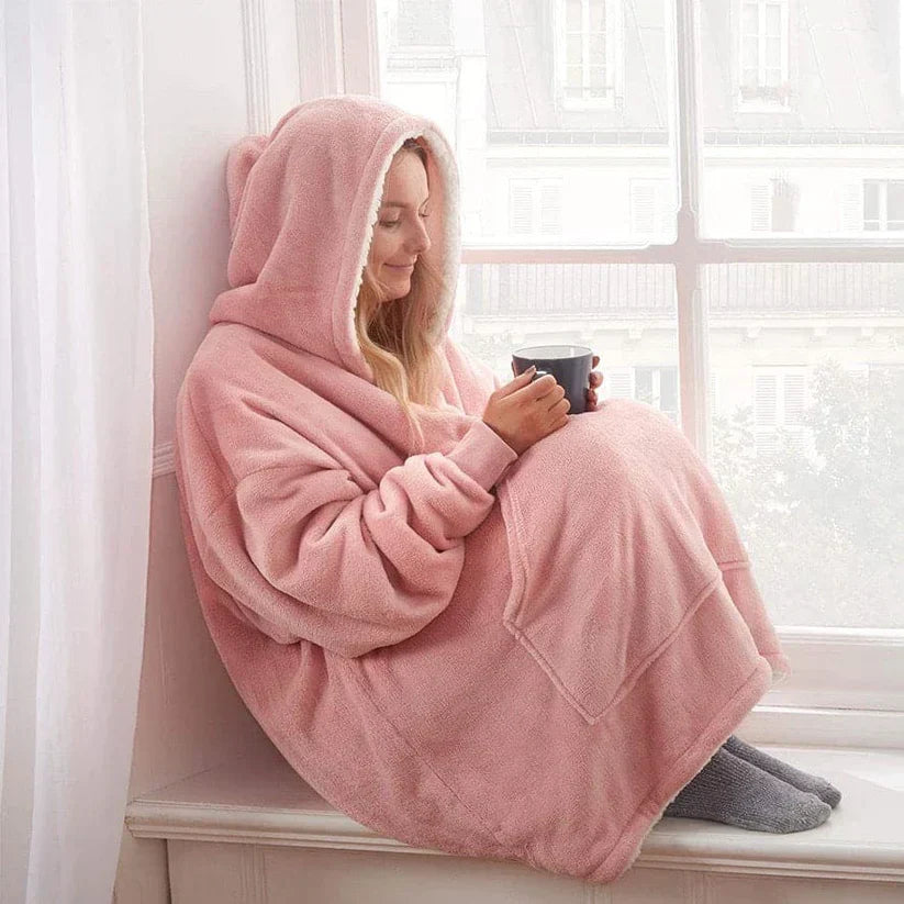Frau in rosa Kuscheldecke mit Kapuze, sitzt am Fenster, hält Tasse, gemütlich, warm, Winter.