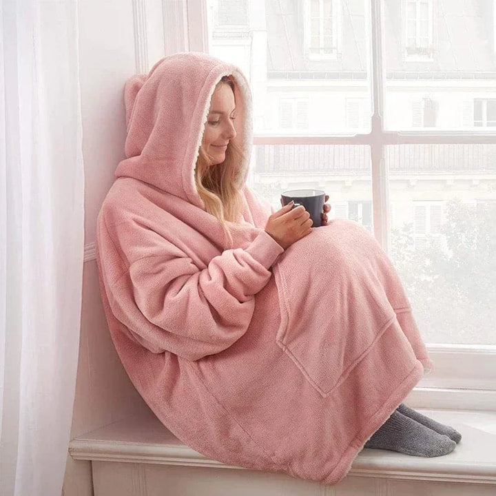 Frau in rosa Kuscheldecke mit Kapuze, sitzt am Fenster, hält Tasse, gemütlich, warm, Winter.