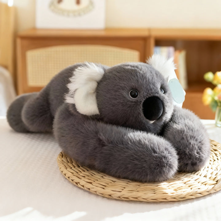 Šedá plyšová koala s bílýma ušima a velkým černým čenichem leží na slaměné podložce s dlouhými pažemi. 16