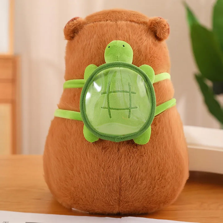 Braunes Capybara-Plüschtier mit grünem Schildkröten-Rucksack auf Holztisch.