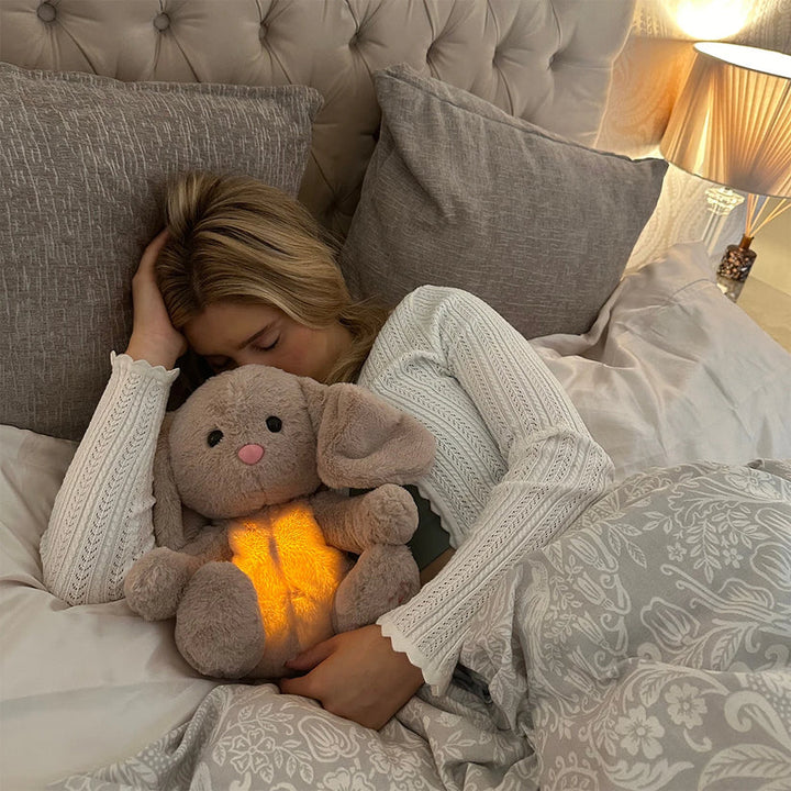 Frau schläft mit leuchtendem Plüschhase im Bett, graue Bettwäsche, gemütliche Schlafzimmerbeleuchtung.