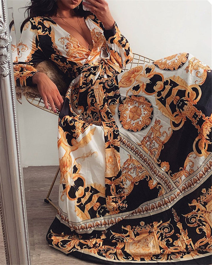 Frau in eleganter, langärmeliger, barockgemusterter Maxikleid in Schwarz und Gold auf Stuhl.