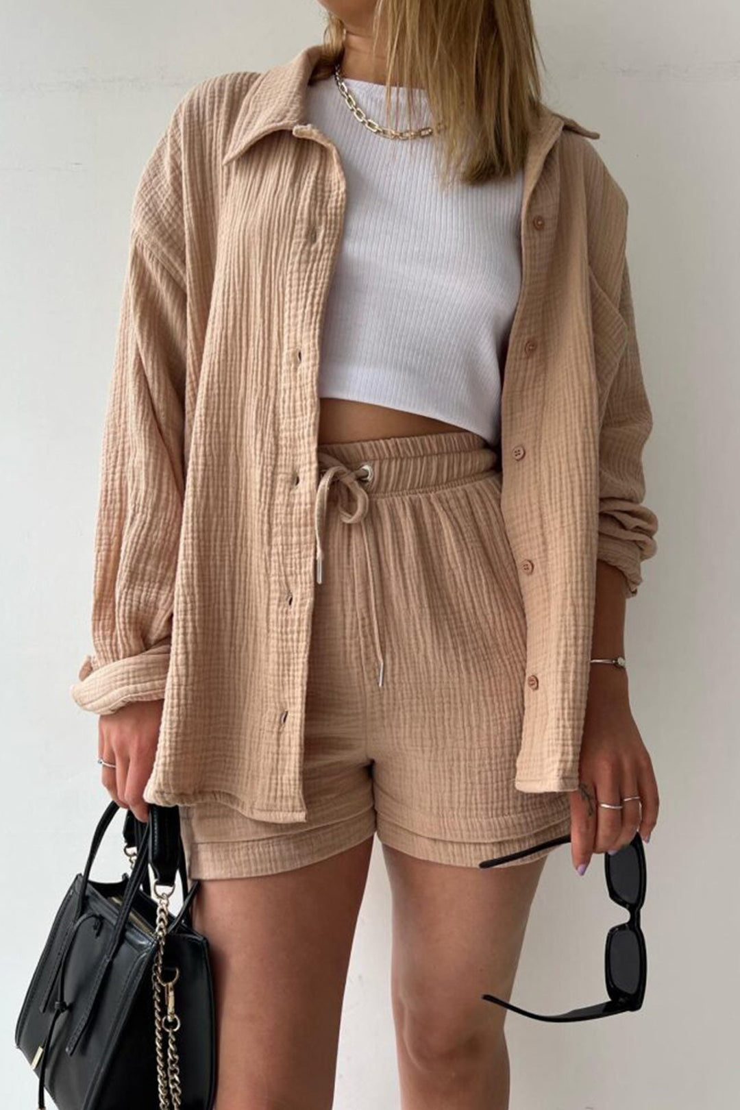 Frau in beigem Langarmhemd und Shorts, weißes Top, schwarze Tasche, Sonnenbrille in der Hand.