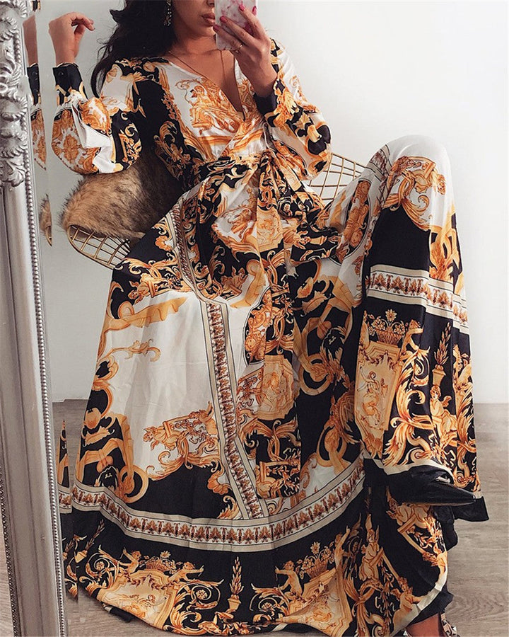 Frau in langem, gemustertem Maxikleid mit barockem Design, Schwarz-Gold-Weiß, sitzt auf Stuhl.