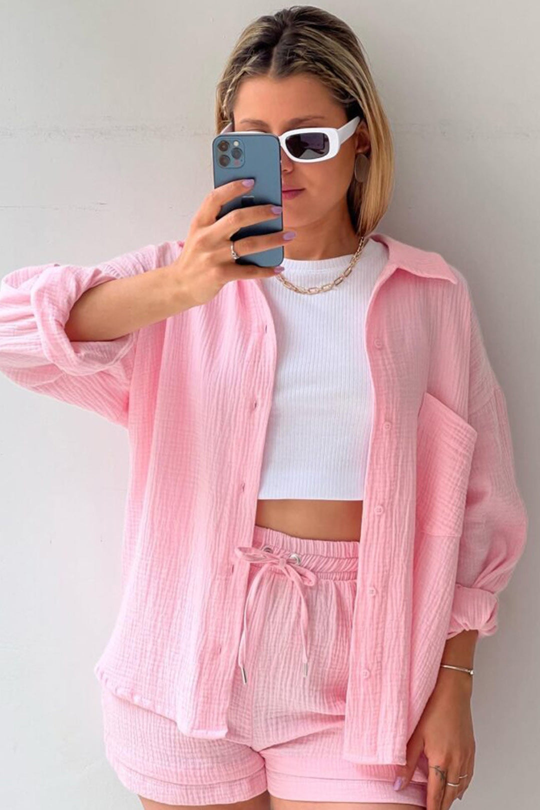 Frau in rosa Langarmhemd und Shorts, weißes Top, Sonnenbrille, Smartphone, modisch.