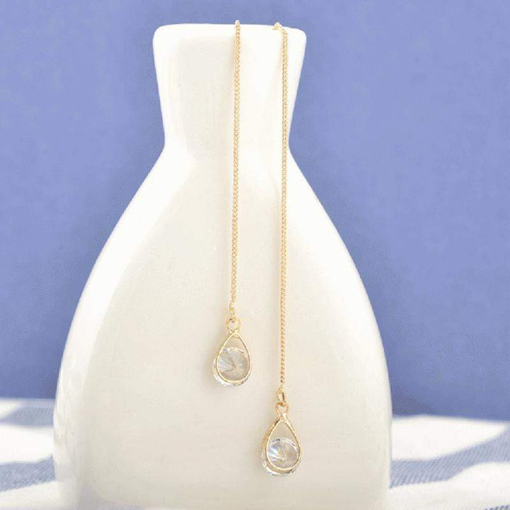Lange goldene Tropfen-Ohrringe mit Strasssteinen auf weißer Vase vor blauem Hintergrund.
