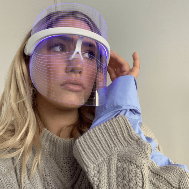 Frau mit LED-Gesichtsmaske, blaues Licht, grauer Pullover, Hautpflege, Anti-Aging-Behandlung.