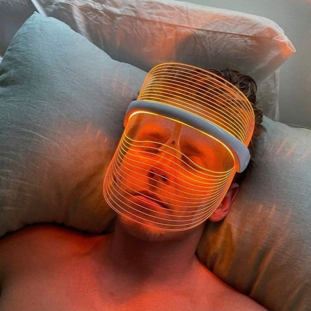 Mann mit LED-Lichttherapie-Maske, orange, auf Kissen liegend, Hautpflege, Entspannung.