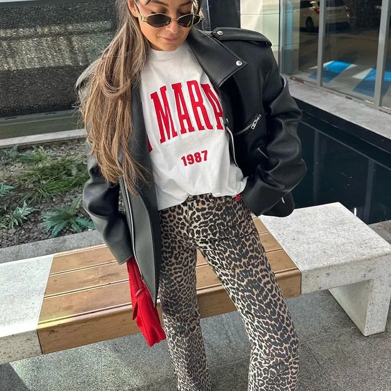 Frau in schwarzer Lederjacke, weißem T-Shirt mit rotem Aufdruck und Leopardenhose, Sonnenbrille.