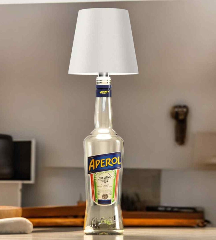 Bílá lampička nasazená na čiré lahvi aperitivu, stojí na kuchyňské lince ve světlém interiéru. 3