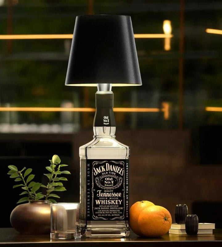 Černá lampička s kovovým hrdlem nasazená na hranaté lahvi whisky, vedle sklenice, pomeranče a rostliny ve váze. 2