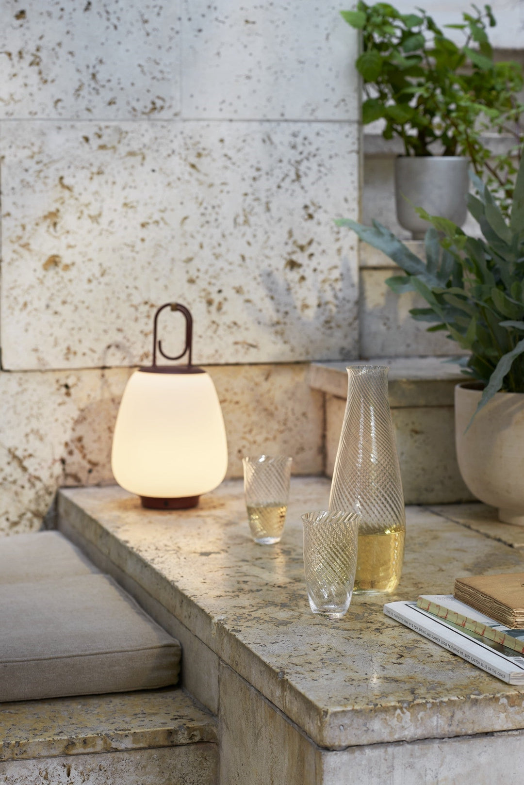 Leuchtende tragbare Lampe auf Steintreppe, umgeben von Pflanzen und Gläsern, moderne Außenbeleuchtung.