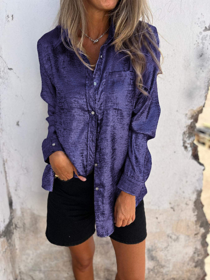 Frau in lila Satinbluse und schwarzer Shorts vor weißer Wand.