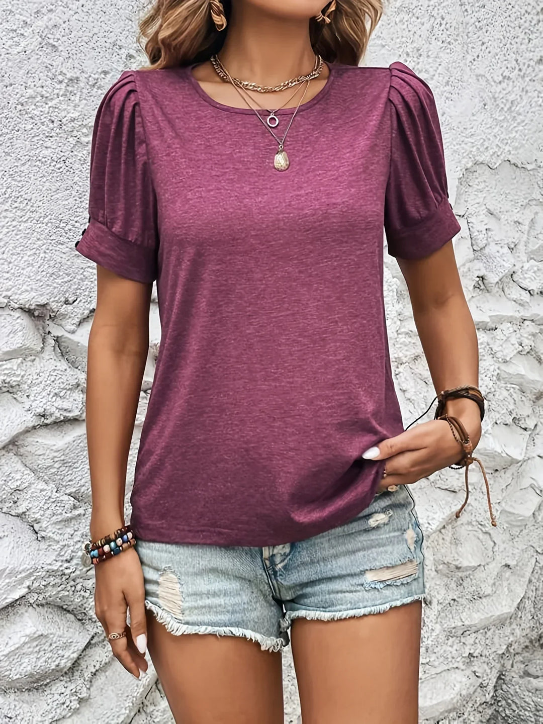 Frau in lila T-Shirt mit Puffärmeln und Jeansshorts vor weißer Wand.