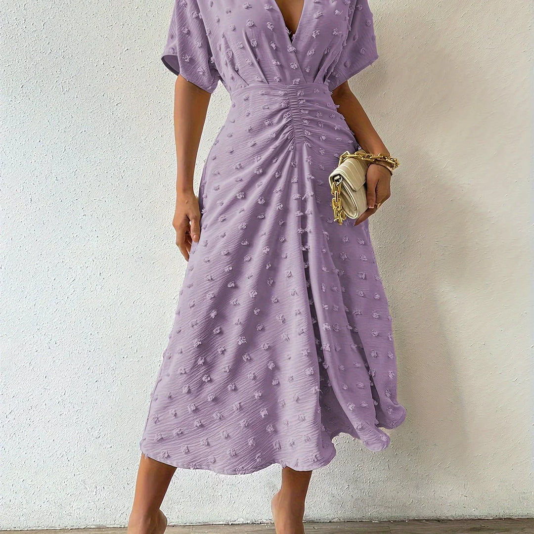 Frau in lila Sommerkleid mit V-Ausschnitt, Raffung, hält beigen Clutch, modisch elegant.