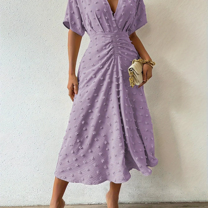 Frau in lila Sommerkleid mit V-Ausschnitt, Raffung, hält beigen Clutch, modisch elegant.