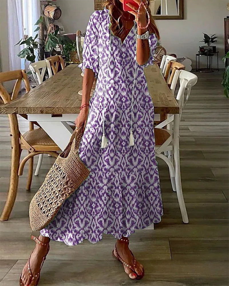 Frau in lila-weißem Maxikleid mit Ikat-Muster, Strohtasche, Sandalen, Esszimmer im Hintergrund.