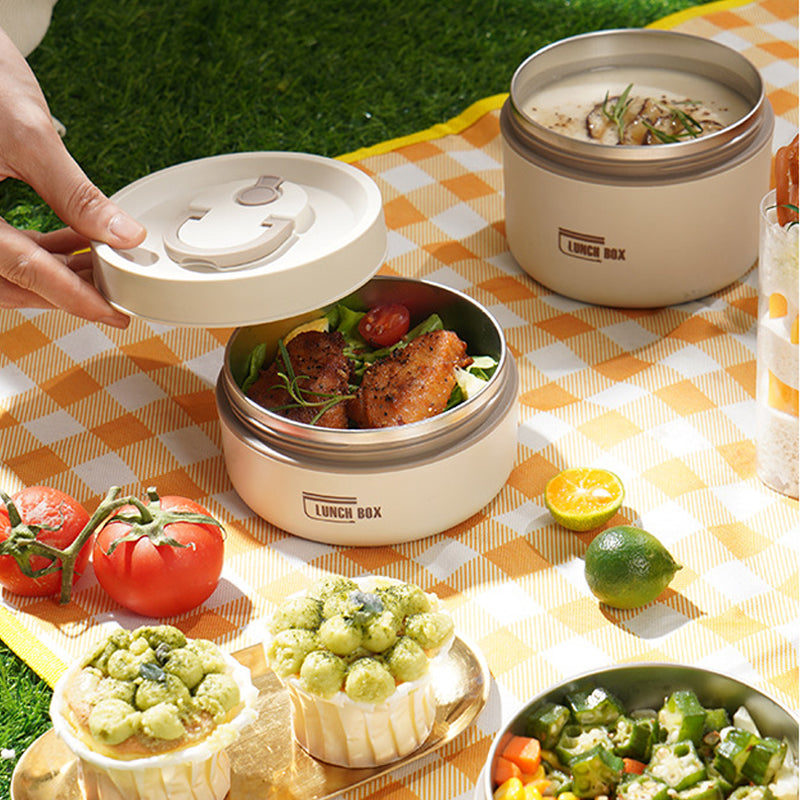 Zwei runde Lunchboxen aus Edelstahl auf Picknickdecke mit Salat und Tomaten.