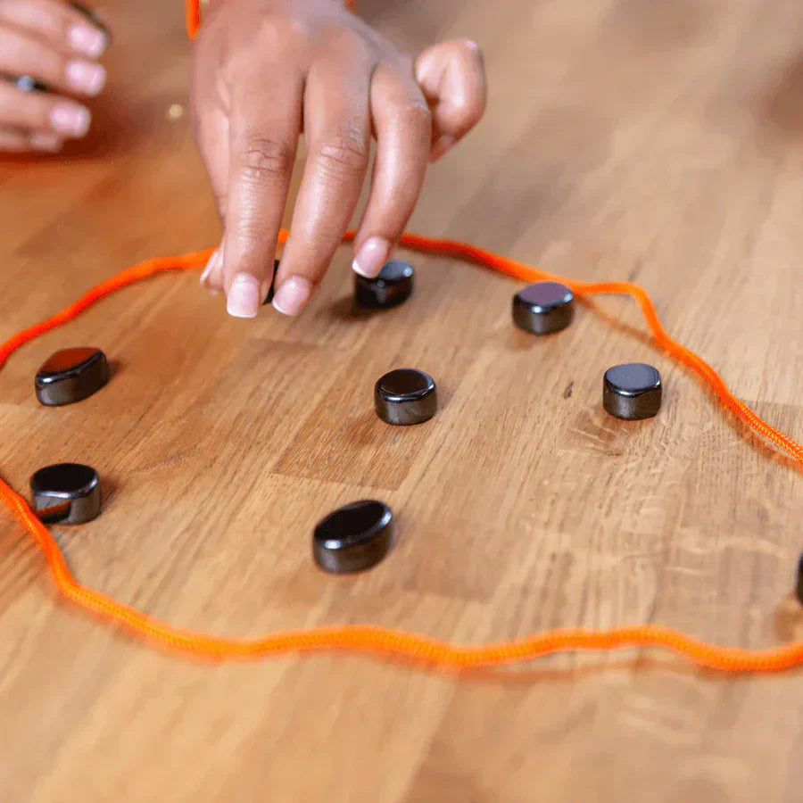 Hand platziert magnetische Steine auf Holzbrett, umgeben von orangefarbener Schnur.