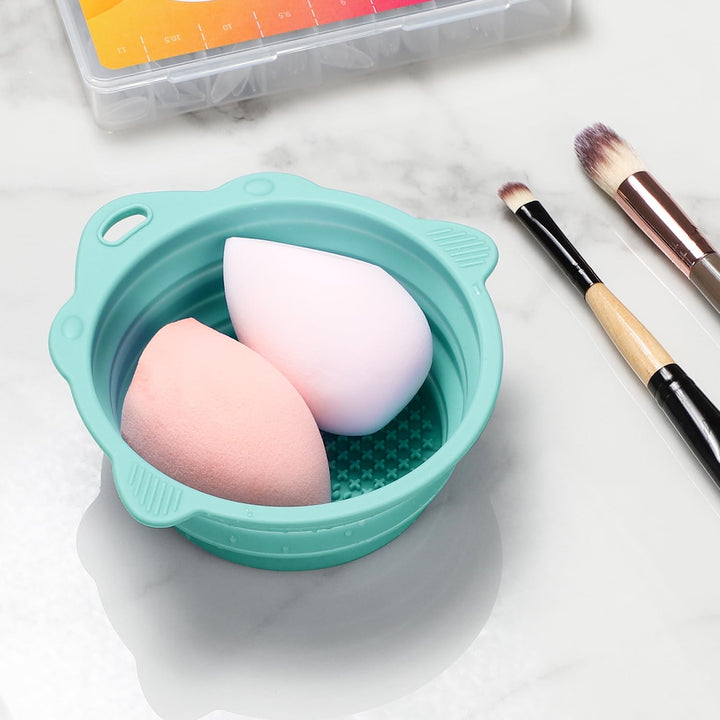 Zwei Make-up-Schwämme in türkisfarbener Reinigungsbox auf Marmoroberfläche, mit Pinseln.