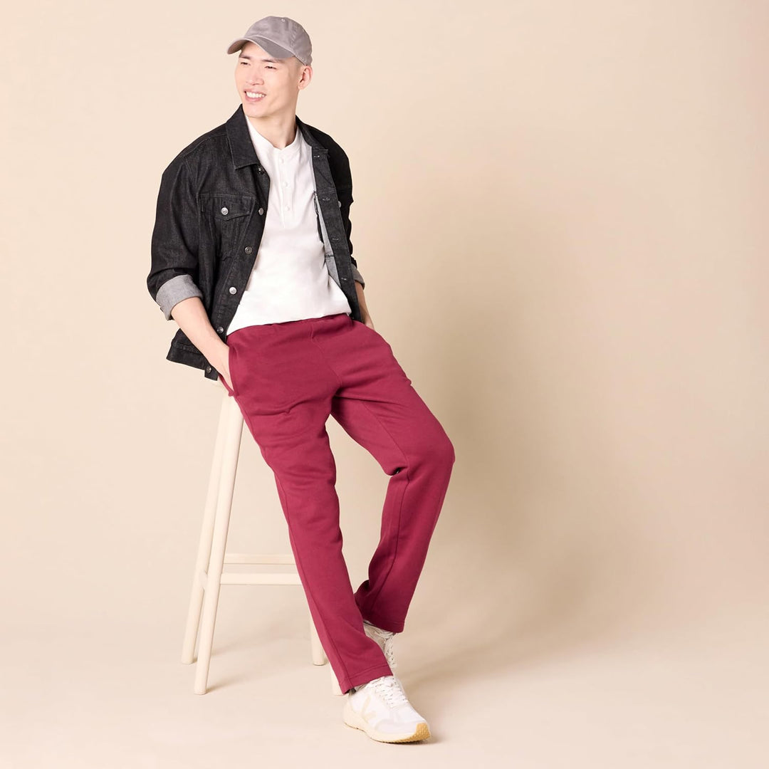 Mann in roter Hose, weißem Shirt, schwarzer Jacke und grauer Kappe auf Hocker, lächelnd.