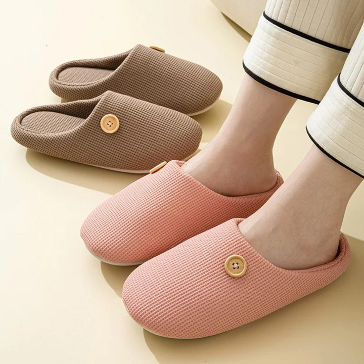 Rosa und braune Memory-Foam-Hausschuhe mit Knopfdetail auf beigem Hintergrund.