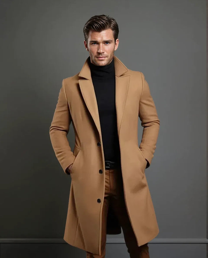 Velbloudí kabát na modelovi v celé postavě, s černým rolákem, páskem a hnědými kalhotami, ruce v kapsách, šedé pozadí. 5