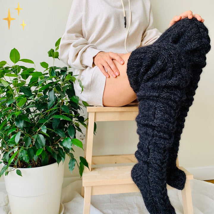 Frau in cremefarbenem Hoodie mit schwarzen Strick-Overknee-Socken auf Holzstuhl neben Pflanze.