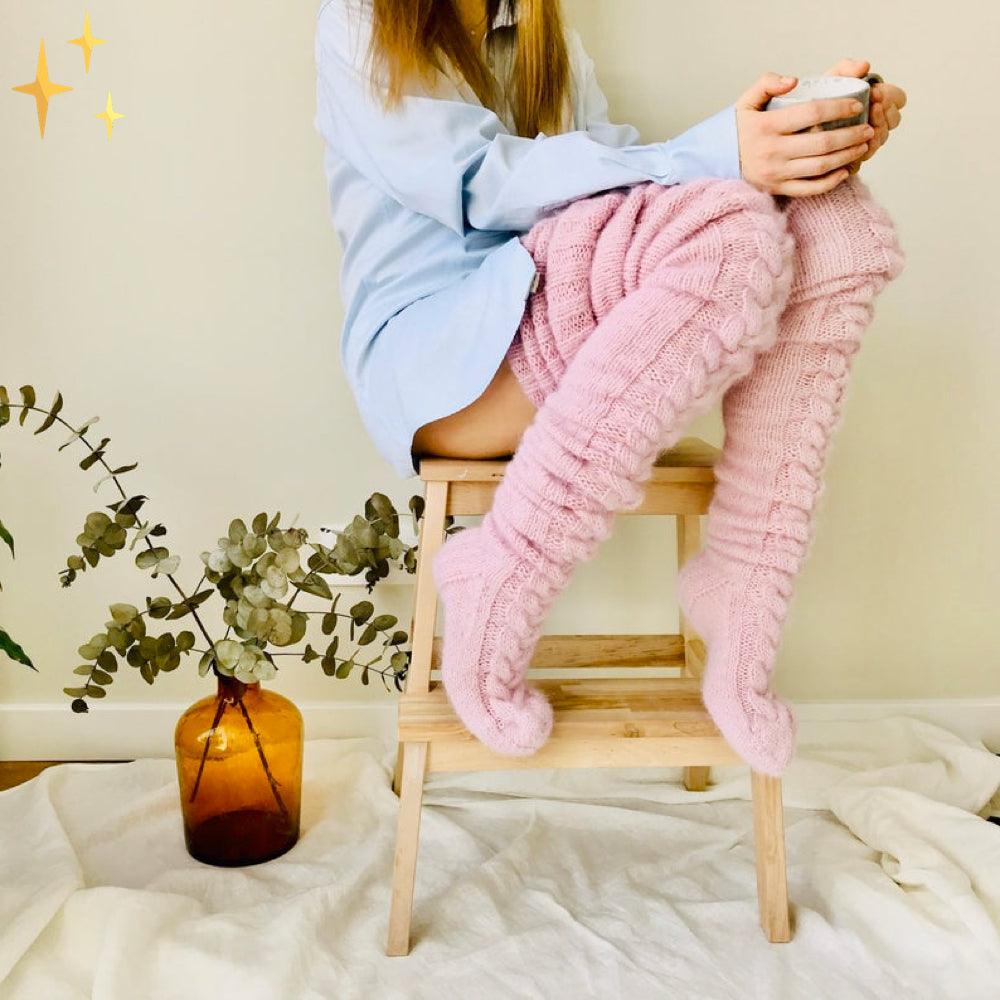 Frau in rosa Strick-Overknee-Socken auf Hocker, gemütlich, mit Tasse, Eukalyptus-Deko.