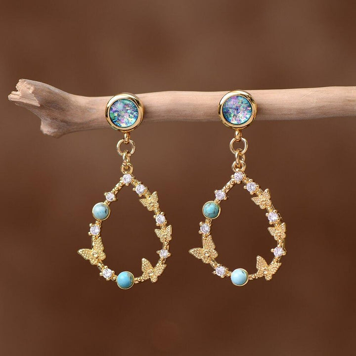 Goldene Ohrringe mit Opal, Zirkonia und Schmetterlingsdesign, hängend an Ast.