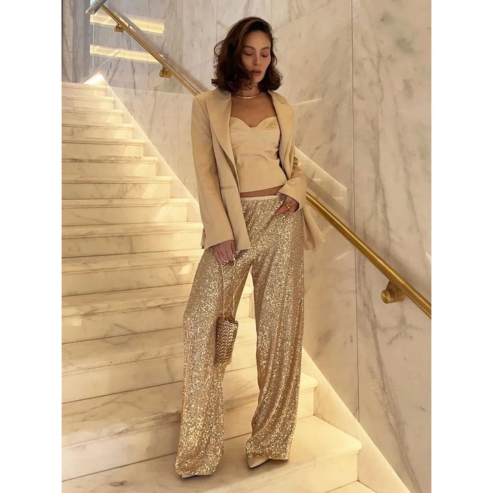 Frau in goldener Paillettenhose und Blazer auf Marmortreppe, elegantes Abendoutfit.