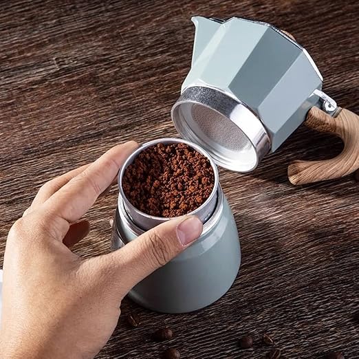 Grüner Espressokocher aus Metall auf Holztisch, Hand füllt Kaffeepulver ein.
