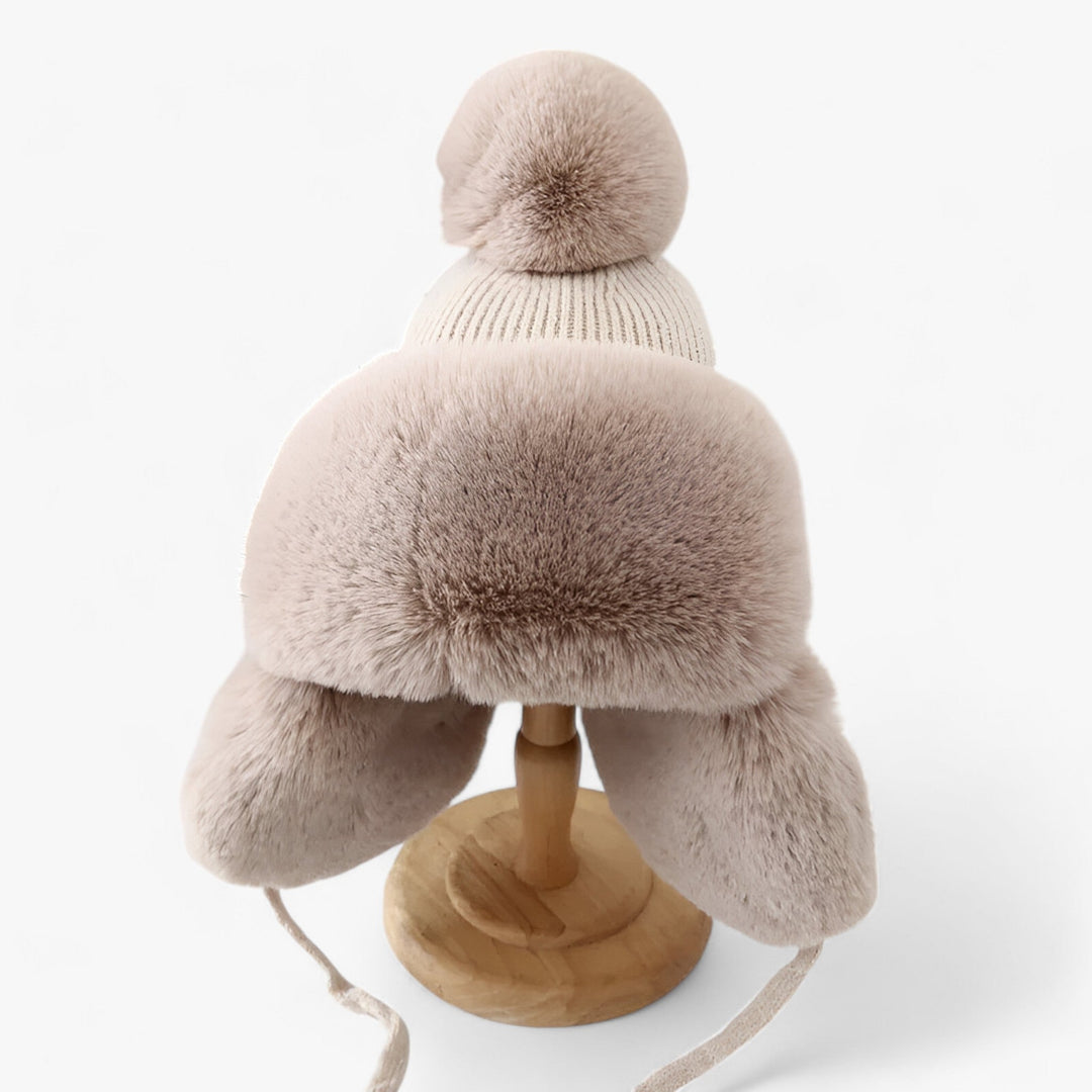 Beige Wintermütze mit Kunstfell, Bommel und Ohrenklappen auf Holzständer, warm und stilvoll.