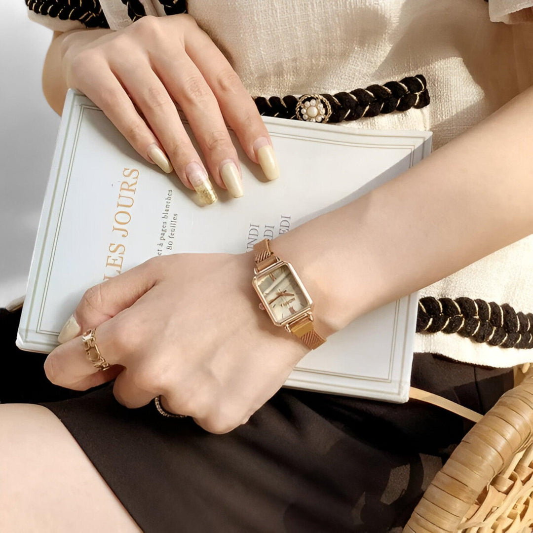 Frau mit goldener Armbanduhr, rechteckiges Zifferblatt, hält ein Buch, elegante Kleidung.