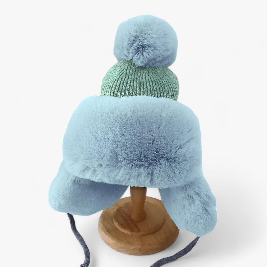 Blaue Wintermütze mit Kunstfell, Bommel und Ohrenklappen auf Holzständer, warm und modisch.